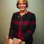 A smiling woman with short blonde hair in a black and burgundy striped jacket, holding a tan leather clutch.