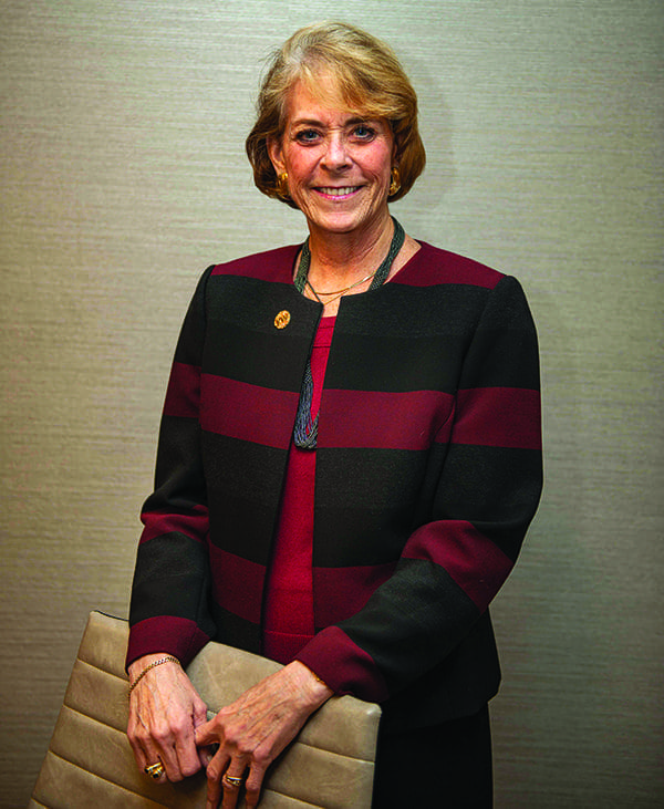 A smiling woman with short blonde hair in a black and burgundy striped jacket, holding a tan leather clutch.