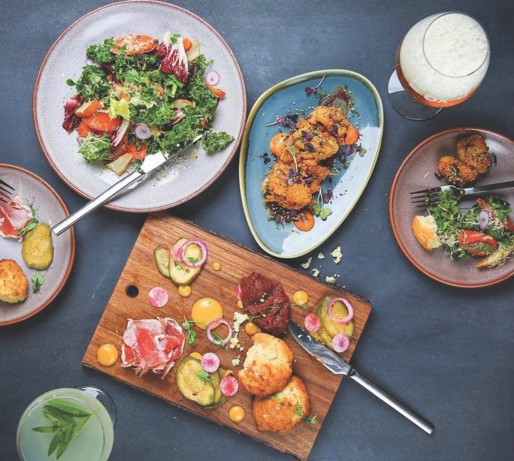 Overhead view of shared plates on a dark table: kale salad, fried cauliflower, charcuterie board, biscuits, and cocktails.