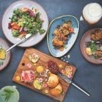 Overhead view of shared plates on a dark table: kale salad, fried cauliflower, charcuterie board, biscuits, and cocktails.