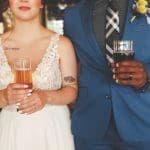 A bride in a white lace dress and a groom in a blue suit hold beers at a brewery.