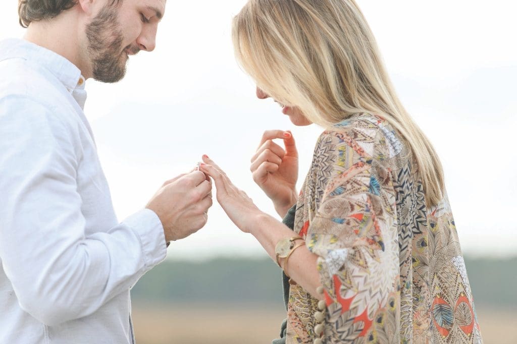 A bearded man in a white shirt slides a ring onto a smiling woman's finger in an open field.