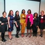 Seven women in dresses and suits pose side by side at an indoor event, with name badges, in front of a projection screen.