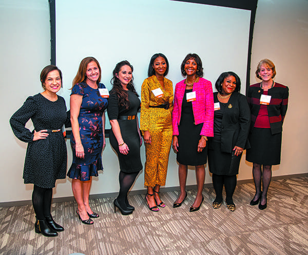 Seven women in dresses and suits pose side by side at an indoor event, with name badges, in front of a projection screen.