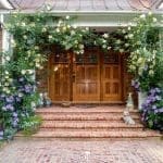 A cedar-shingled house entrance framed by yellow climbing roses and purple clematis, with brick steps and garden statuary.