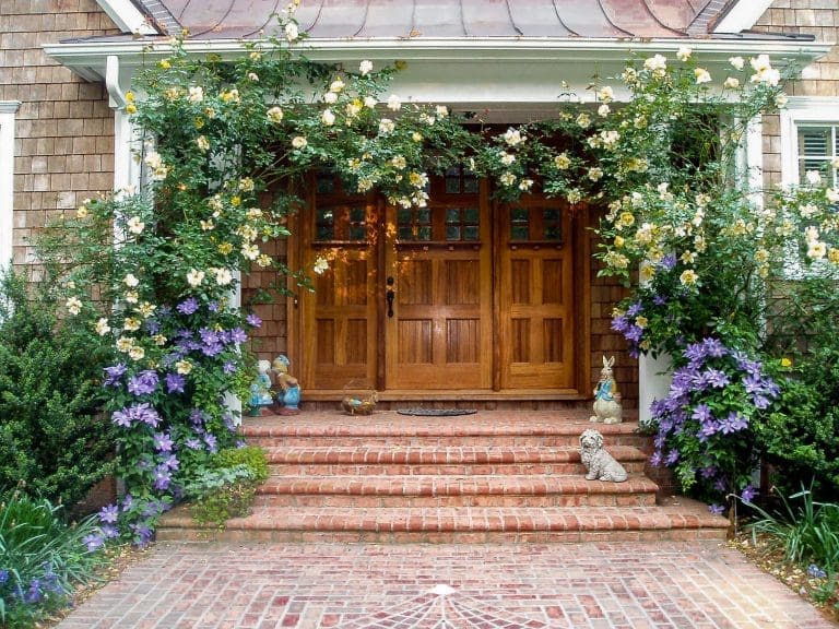 A cedar-shingled house entrance framed by yellow climbing roses and purple clematis, with brick steps and garden statuary.