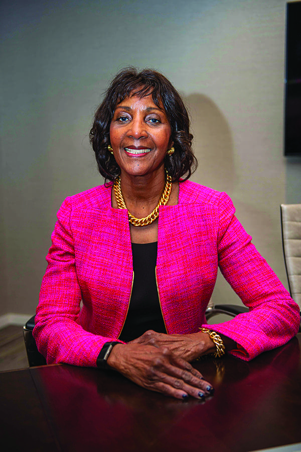 A smiling woman in a bright pink tweed jacket and gold chain necklace sits at a dark wooden conference table.