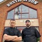 Two chefs in black jackets stand before a brick building with "The Rustic Spoon" sign above it.