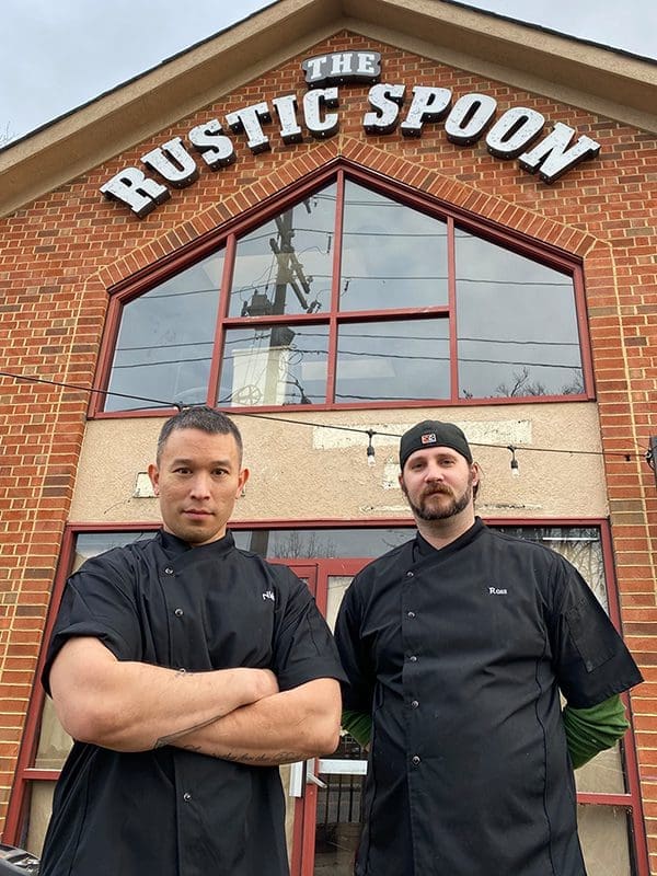 Two chefs in black jackets stand before a brick building with "The Rustic Spoon" sign above it.
