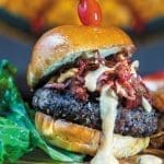 A burger on a brioche bun with crispy bacon, creamy white sauce, a cherry pick, lettuce and fries.