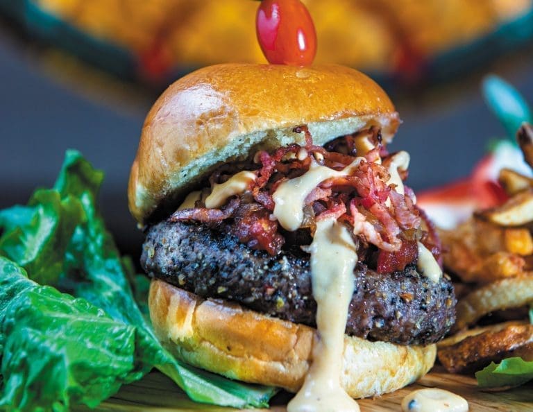A burger on a brioche bun with crispy bacon, creamy white sauce, a cherry pick, lettuce and fries.