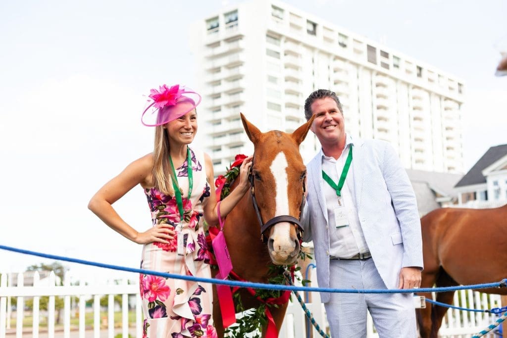 A smiling woman in a pink hat and floral dress and a man in a seersucker suit stand beside a chestnut horse with roses.