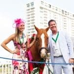 A smiling woman in a pink hat and floral dress and a man in a seersucker suit stand beside a chestnut horse with roses.