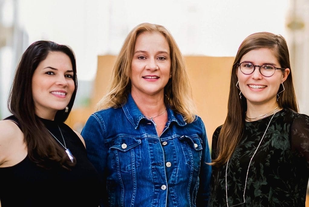 Three smiling women posing side by side; the woman in the middle wears a denim jacket and the others wear dark tops.
