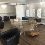A bright salon interior with black styling chairs, beige sofas, a "Hello Gorgeous" pillow, and white barn-style mirrors.