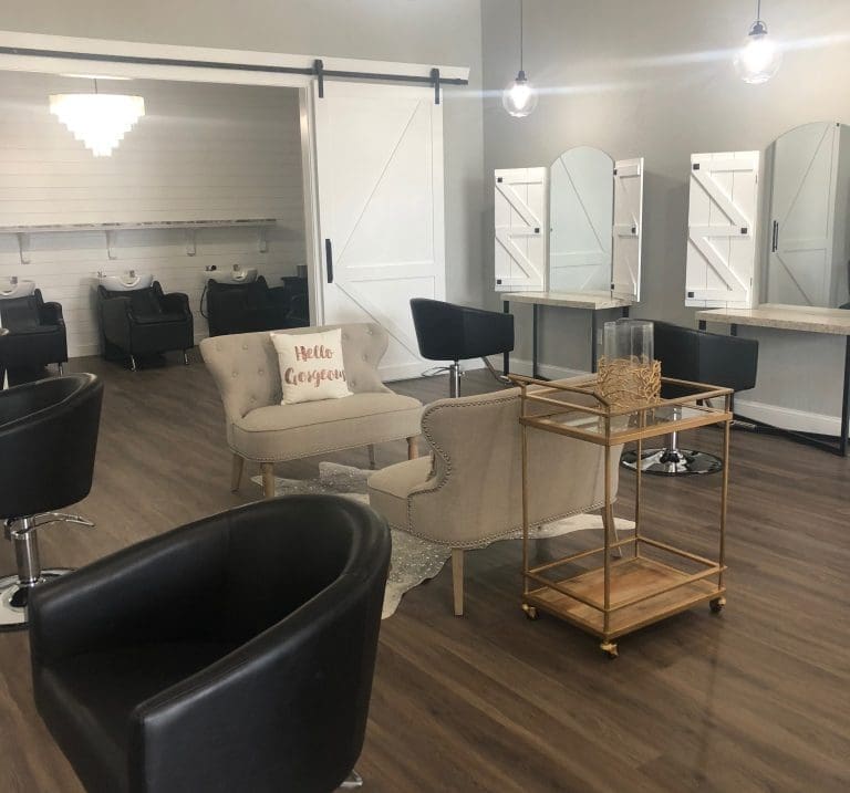 A bright salon interior with black styling chairs, beige sofas, a "Hello Gorgeous" pillow, and white barn-style mirrors.