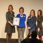Four women stand on a stage, one holding a framed "Giving Back Awards" certificate, as a person photographs them.