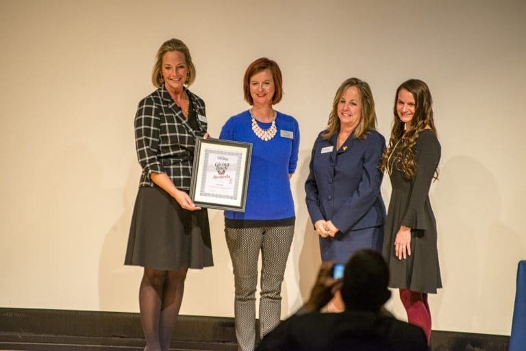 Four women stand on a stage, one holding a framed "Giving Back Awards" certificate, as a person photographs them.