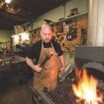 A bald bearded blacksmith in a leather apron holds a tool beside a roaring forge fire in a workshop.