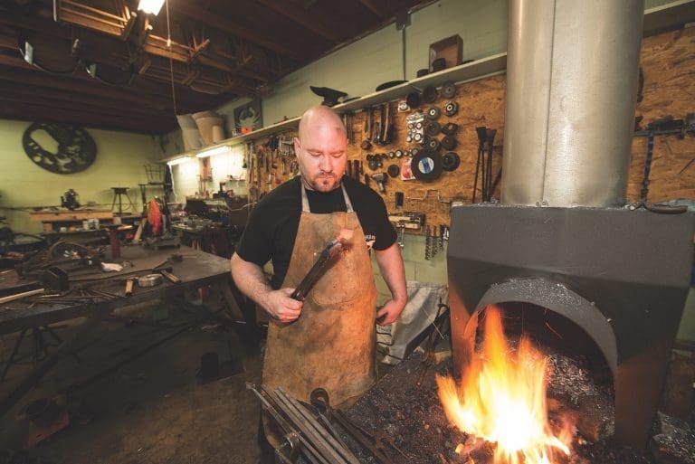 A bald bearded blacksmith in a leather apron holds a tool beside a roaring forge fire in a workshop.