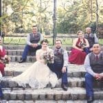 Bridal party of a bride in a lace gown and groomsmen in gray vests and burgundy ties posing on stone steps before an iron gate.