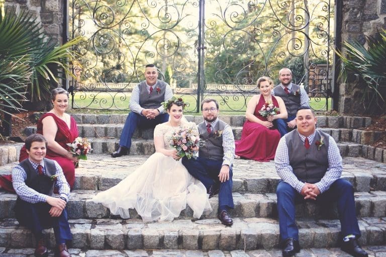 Bridal party of a bride in a lace gown and groomsmen in gray vests and burgundy ties posing on stone steps before an iron gate.