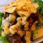 A burger topped with melted cheese, crispy tortilla strips, and green chiles, served on a bed of leafy lettuce.
