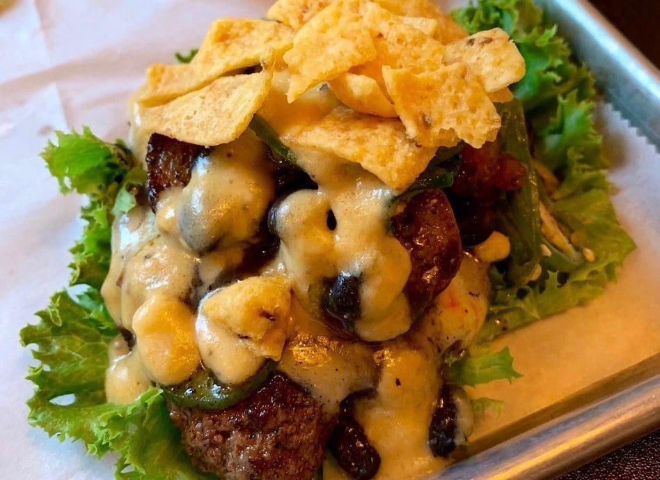 A burger topped with melted cheese, crispy tortilla strips, and green chiles, served on a bed of leafy lettuce.