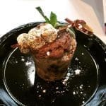 A cookie-dough dessert cup topped with toasted marshmallows, chocolate frosting, a chocolate curl, and fresh mint on a black plate.