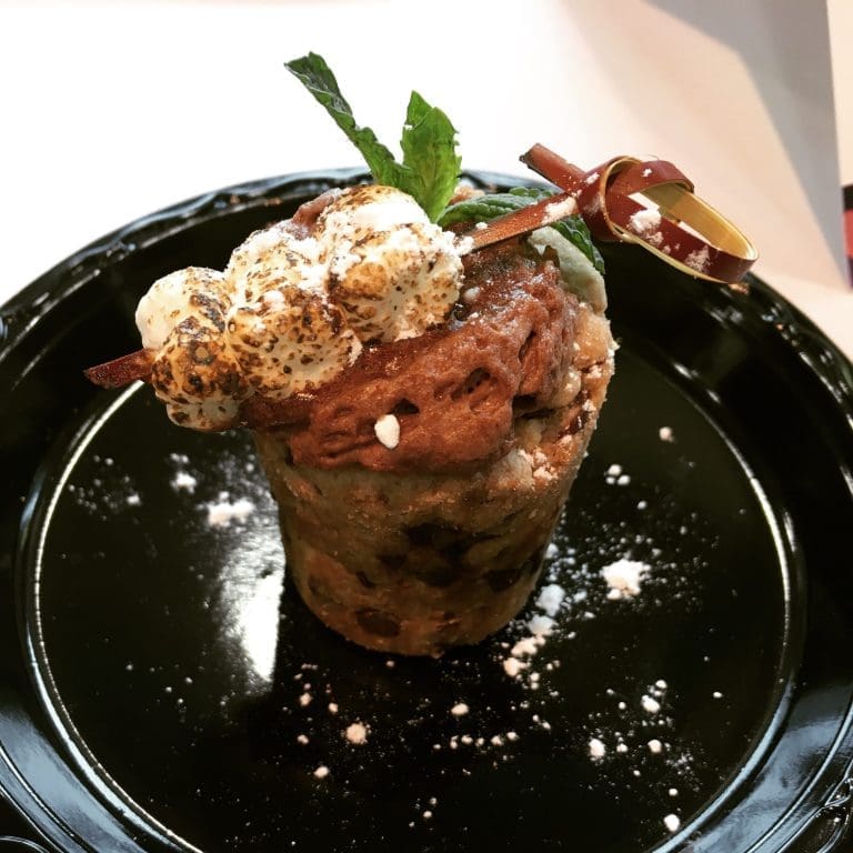 A cookie-dough dessert cup topped with toasted marshmallows, chocolate frosting, a chocolate curl, and fresh mint on a black plate.