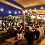 Evening crowd gathers under string lights at long wooden picnic tables in an outdoor beer garden.