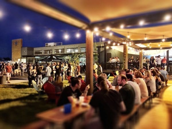 Evening crowd gathers under string lights at long wooden picnic tables in an outdoor beer garden.