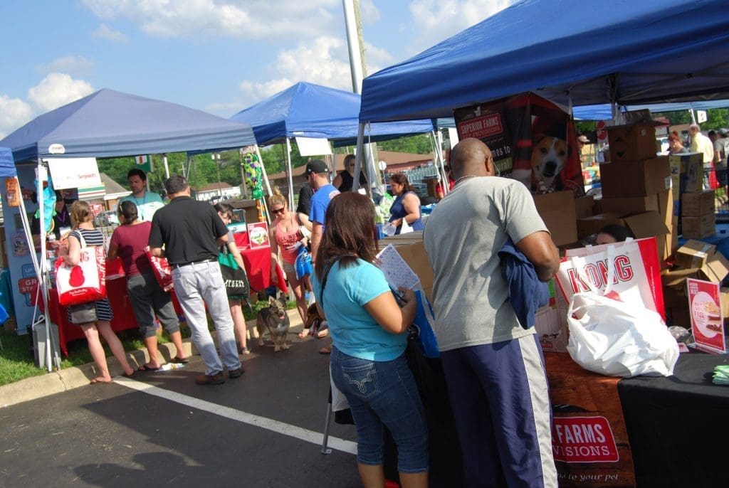 Shoppers browse pet vendor tents under blue canopies, including a Kong booth and Supreme Farms pet food display, at an outdoor event.