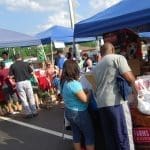 Shoppers browse pet vendor tents under blue canopies, including a Kong booth and Supreme Farms pet food display, at an outdoor event.
