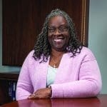 A smiling woman with gray locs and glasses wears a lavender cardigan, leaning on a polished wooden desk.