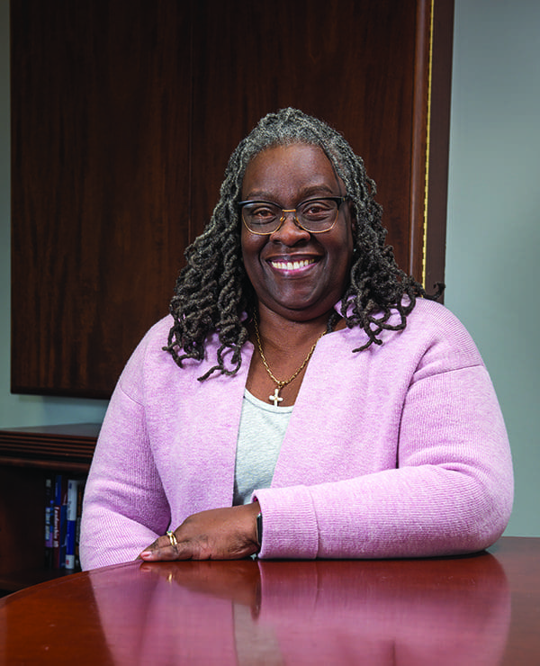 A smiling woman with gray locs and glasses wears a lavender cardigan, leaning on a polished wooden desk.