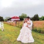 A bride in a blush ruffled gown and a groom in a tan vest embrace in a grassy field beside a white fence and red barn.