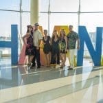 Six people pose in front of large colorful HAVANA letters in a bright waterfront room with glass windows.