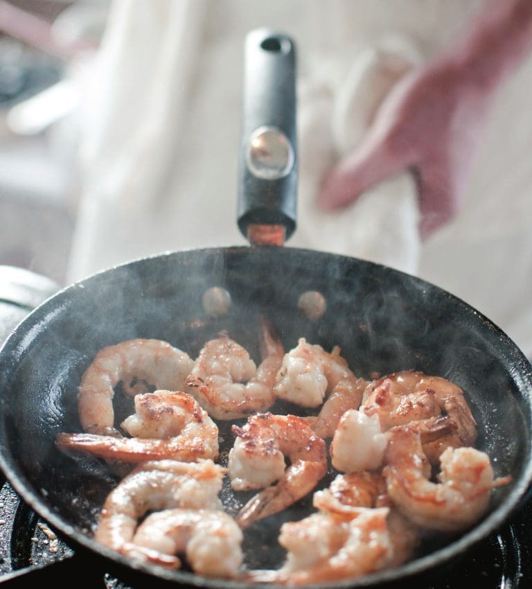 Shrimp sautéing in a dark skillet, steam rising, with a cook in a white apron holding the handle.