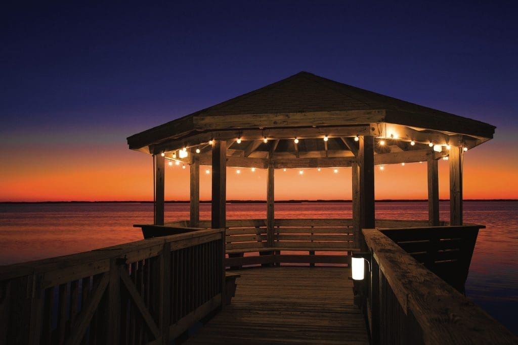 Wooden gazebo with string lights at the end of a pier over calm water at dusk.