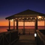Wooden gazebo with string lights at the end of a pier over calm water at dusk.