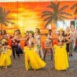A group of dancers in yellow grass skirts and flower leis performs hula in front of a painted tropical sunset backdrop.