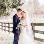 A bride in a white gown and veil and a groom in a navy suit stand foreheads together beside a wooden fence.