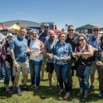 A large group of smiling adults stands outdoors on grass at a sunny festival, holding food plates and cups.