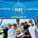 Six men pose under a Bay Local banner, one holding a gold trophy, with a thumbs-up from a man in a Bay Local tag.