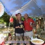 A smiling woman and man stand behind a booth under a blue canopy, holding jars of salsa and selling rows of jars.