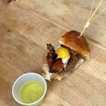 Mini brioche slider with fried egg, bacon and patty on a wooden board, beside a cup of yellow sauce.