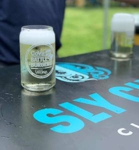 A frothy beer glass branded "CoVa Battle of the Burgers" sits on a black table with blue "SLY CLYDE" lettering.