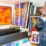A smiling man in a paint-splattered shirt and hat holds the book "Poetry Art n Truth" in a studio of colorful paintings.