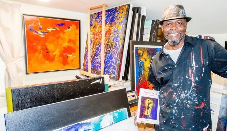 A smiling man in a paint-splattered shirt and hat holds the book "Poetry Art n Truth" in a studio of colorful paintings.